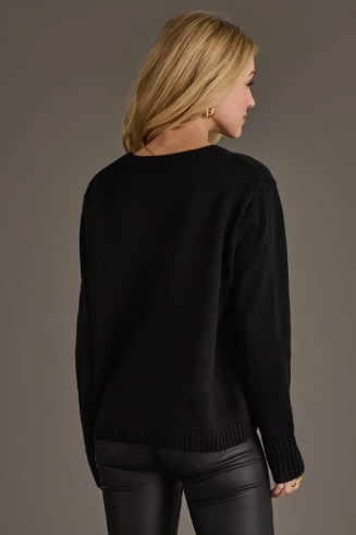 A woman with blonde hair is shown from the back wearing a black knit sweater and black faux-leather pants against a solid dark gray studio background.