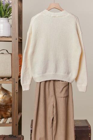 An off-white knit crewneck sweater displayed on a wooden hanger, styled with beige wide-leg trousers in front of a neutral background with rustic wooden shelving.