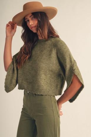 A woman wearing a cropped olive green knit sweater with wide draped sleeves, matching olive green pants, and a tan wide-brim hat, posed against a plain light beige studio background.