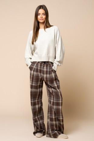 A woman modeling a cozy outfit consisting of a cream-colored crewneck sweatshirt and brown plaid wide-leg pants, standing against a solid beige studio background.