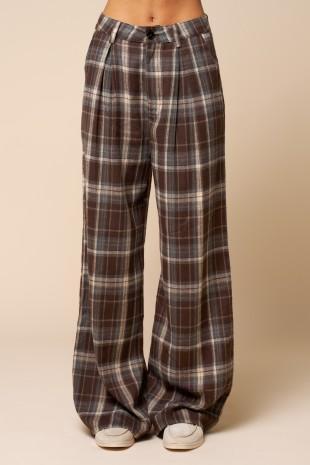 A person wearing high-waisted, wide-leg plaid pants in brown, beige, and gray, paired with white sneakers, shown from the waist down against a solid beige background.