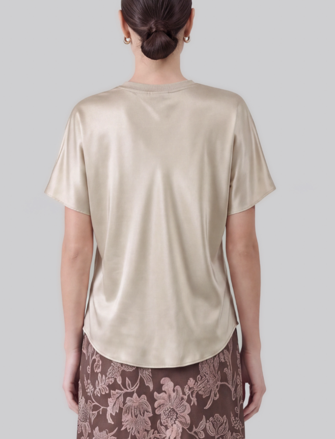 A woman viewed from the back wearing a short-sleeve satin-like top in a light champagne beige color, paired with a brown floral skirt, against a solid light gray background.
