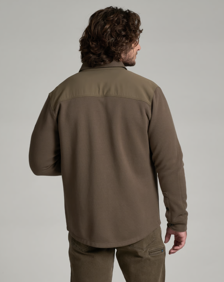 A man viewed from the back wearing a brown Kuhl Relentr long-sleeve fleece pullover with a slightly lighter olive-brown yoke across the shoulders, paired with brown pants, against a solid light gray studio background.