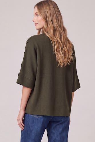 A woman shown from the back wearing an olive green short-sleeve top with rolled cuffs and blue jeans, photographed against a plain light gray studio background.