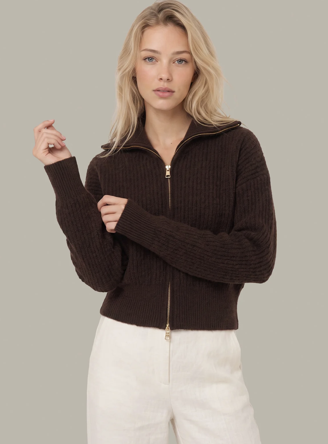 A blonde woman wearing a dark brown ribbed knit 525 America zip-up sweater with a stand collar and white pants, posing against a solid light gray background.