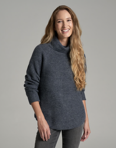 A woman with long blonde hair wearing a dark slate blue Kuhl Sienna ribbed knit turtleneck sweater and gray pants, standing against a solid gray studio background.
