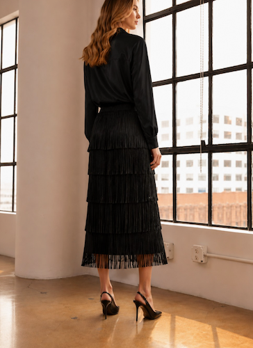 A woman stands with her back to the camera in a black long-sleeve top and a black tiered fringe midi skirt, paired with black high heels, in a bright loft with large industrial windows.