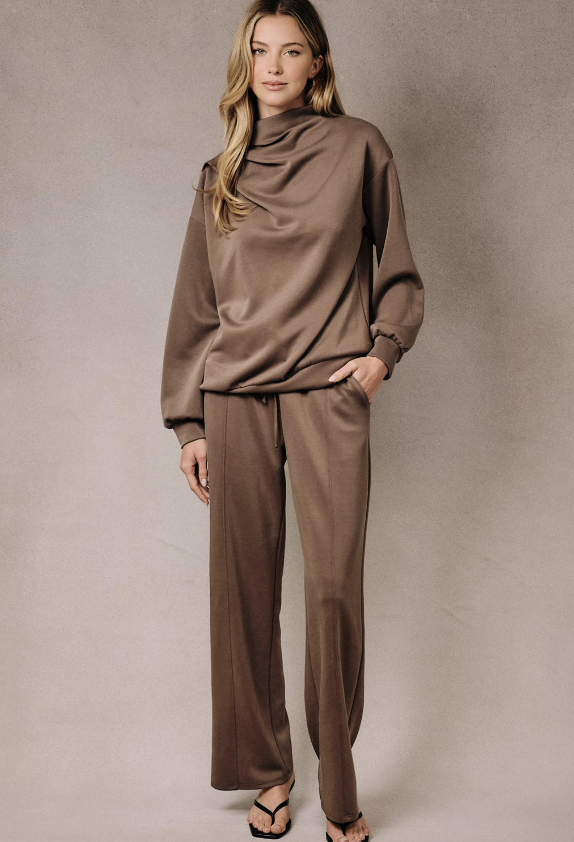A woman modeling a matching two-piece lounge set in a warm taupe-brown color, featuring a draped cowl-neck long-sleeve top and wide-leg pants, set against a neutral textured studio backdrop.