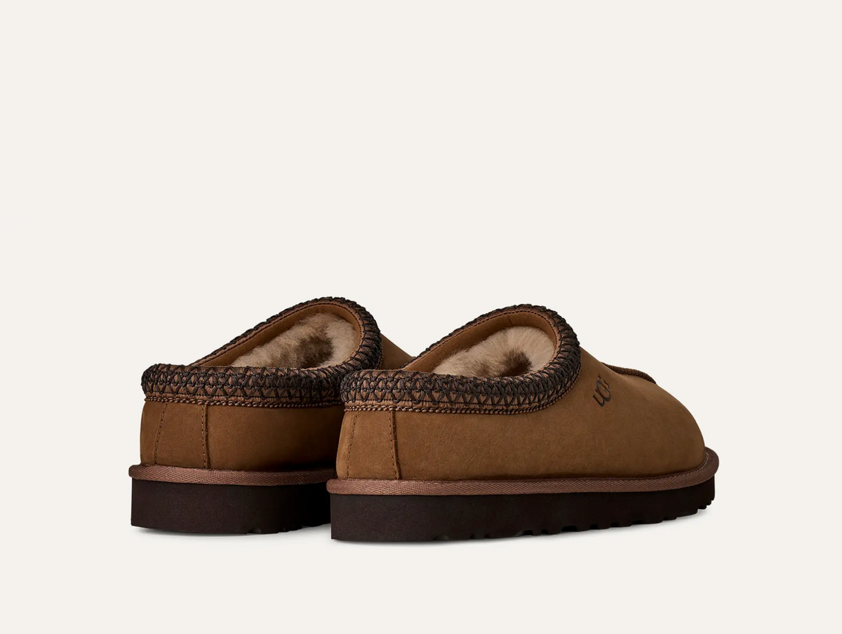 A pair of UGG Men’s Tasman II Nubuck brown suede slip-on slippers with a dark brown braided trim and a soft beige fleece lining, shown from a rear three-quarter angle on a light off-white background.