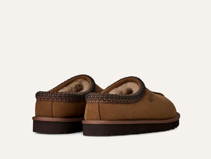 A pair of UGG Men’s Tasman II Nubuck brown suede slip-on slippers with a dark brown braided trim and a soft beige fleece lining, shown from a rear three-quarter angle on a light off-white background.