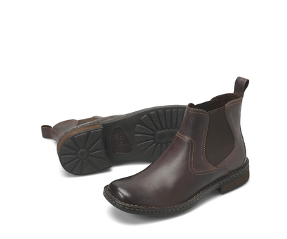 A pair of dark brown leather Chelsea boots, with one boot standing upright to show the side profile and the other lying on its side to display the black rubber sole.