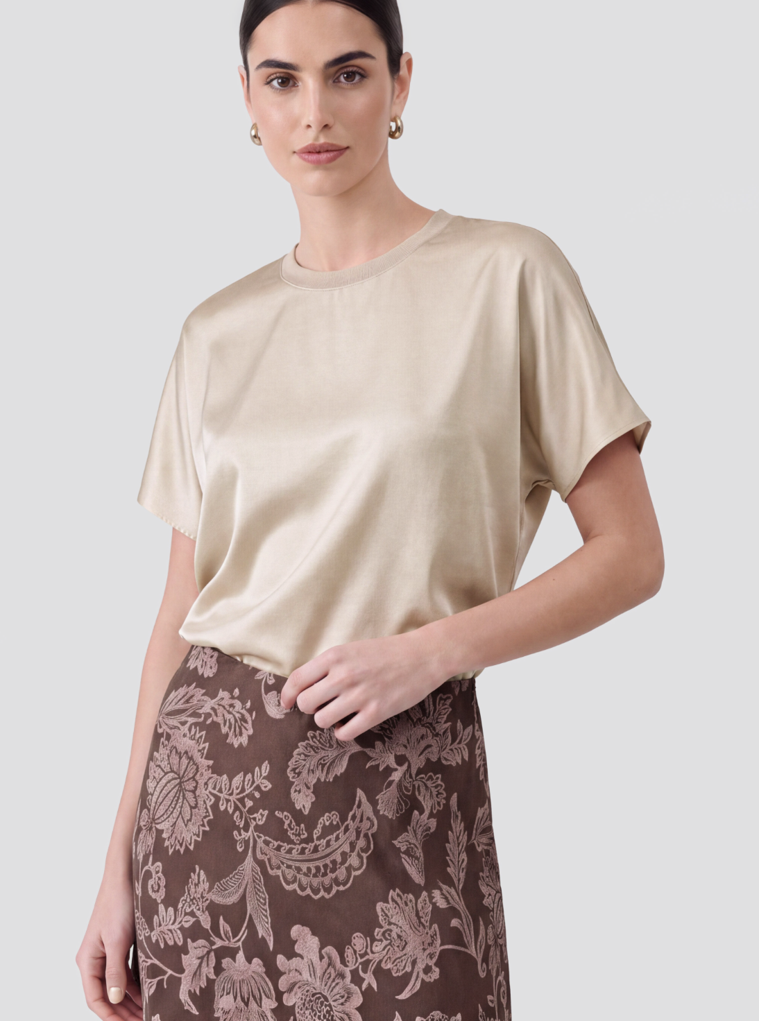 A woman modeling a short-sleeve beige satin top paired with a brown floral print skirt, set against a solid light gray background.