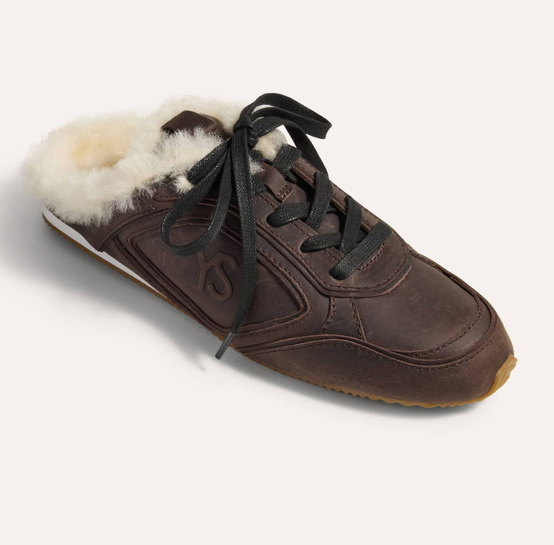 A Yosi Samra dark brown leather slip-on sneaker with a fluffy cream shearling lining and black laces, shown from a three-quarters angle on a light beige background.