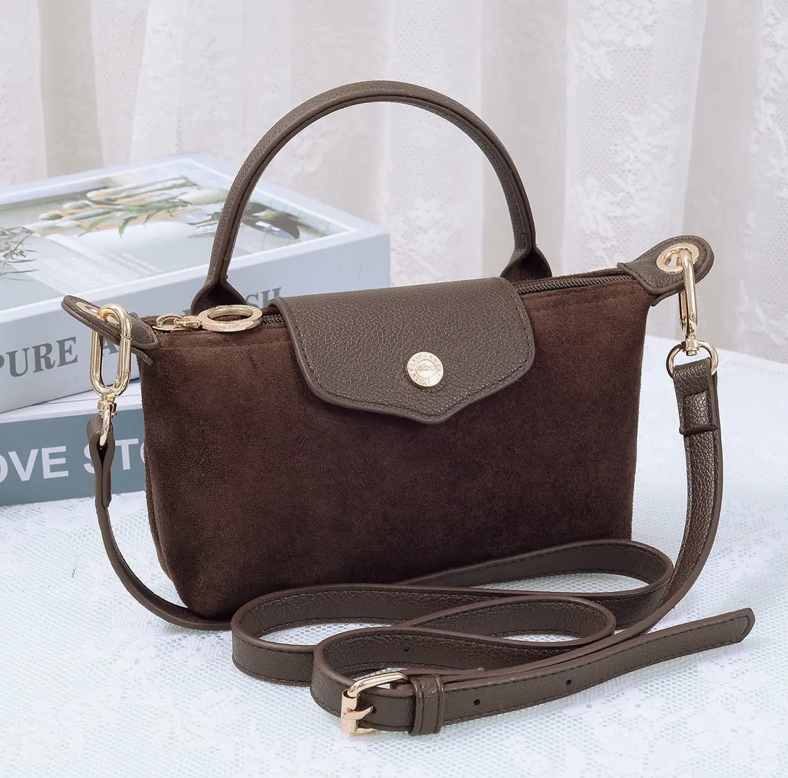 A dark brown suede handbag with a top handle, a fold-over flap secured by a gold-tone button, and a detachable shoulder strap, displayed on a light surface with decorative books in the background.