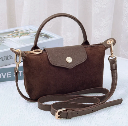 A dark brown suede handbag with a top handle, a fold-over flap secured by a gold-tone button, and a detachable shoulder strap, displayed on a light surface with decorative books in the background.