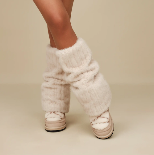 A person wearing fluffy cream UGG faux-fur leg warmers over beige platform sneakers, shown from the knees down against a plain beige studio background.