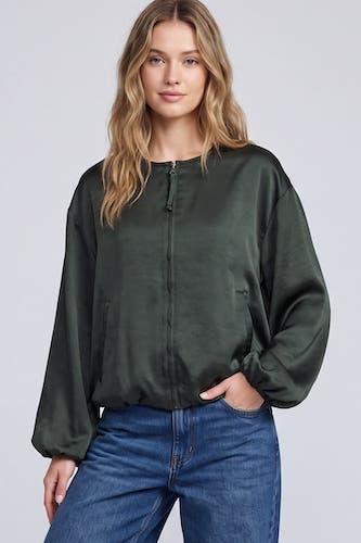 A woman modeling a dark olive green satin zip-up jacket with balloon sleeves, paired with blue jeans, against a light gray studio background.