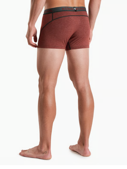 Rear view of a man wearing heathered rust-brown Kuhl boxer briefs with a dark waistband and black trim, standing barefoot against a light gray studio background.