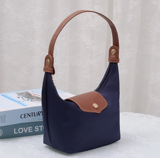 A navy blue nylon shoulder bag with brown faux-leather trim and a gold-tone snap button, displayed on a white textured surface next to a book.