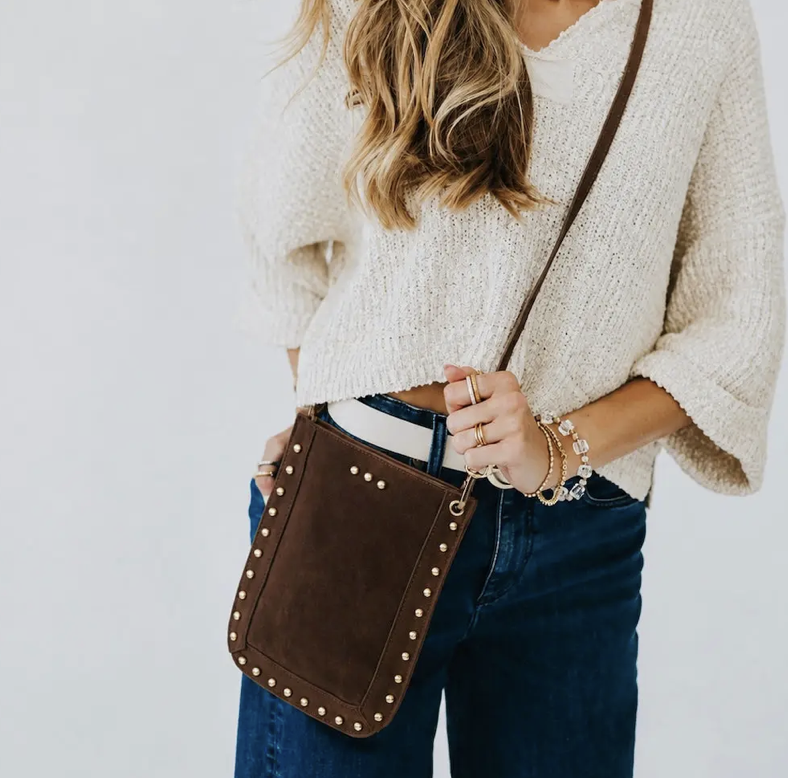 A woman carrying a brown crossbody phone bag with gold-tone studs and a thin brown strap.