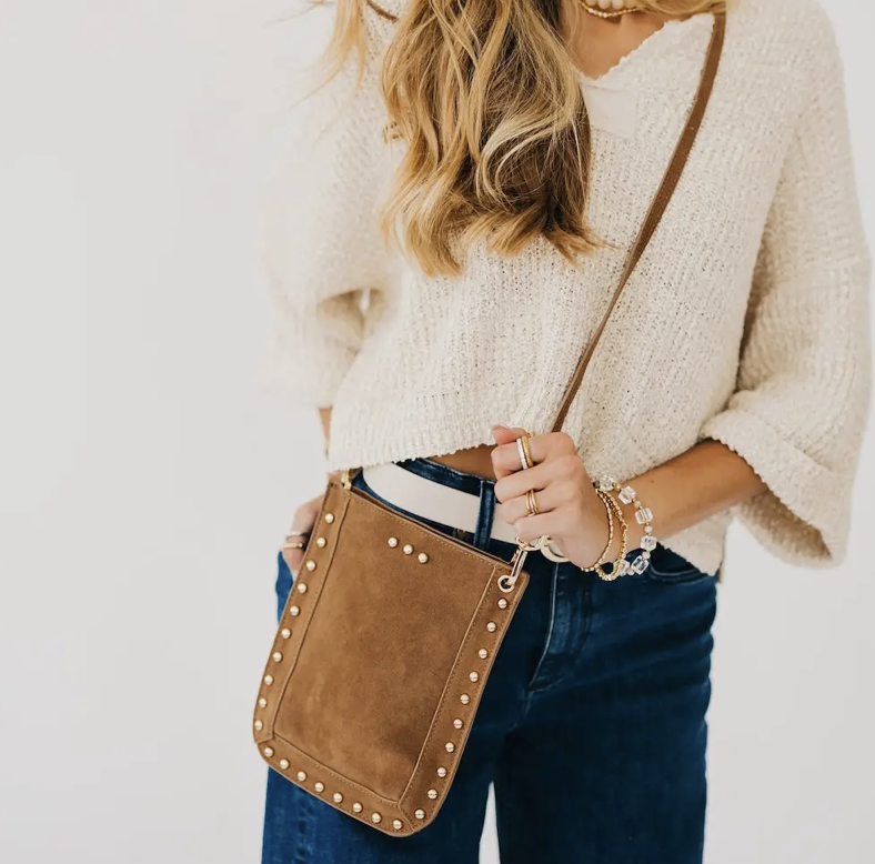 A woman carrying a brown suede crossbody bag with gold-tone studs and a strap.