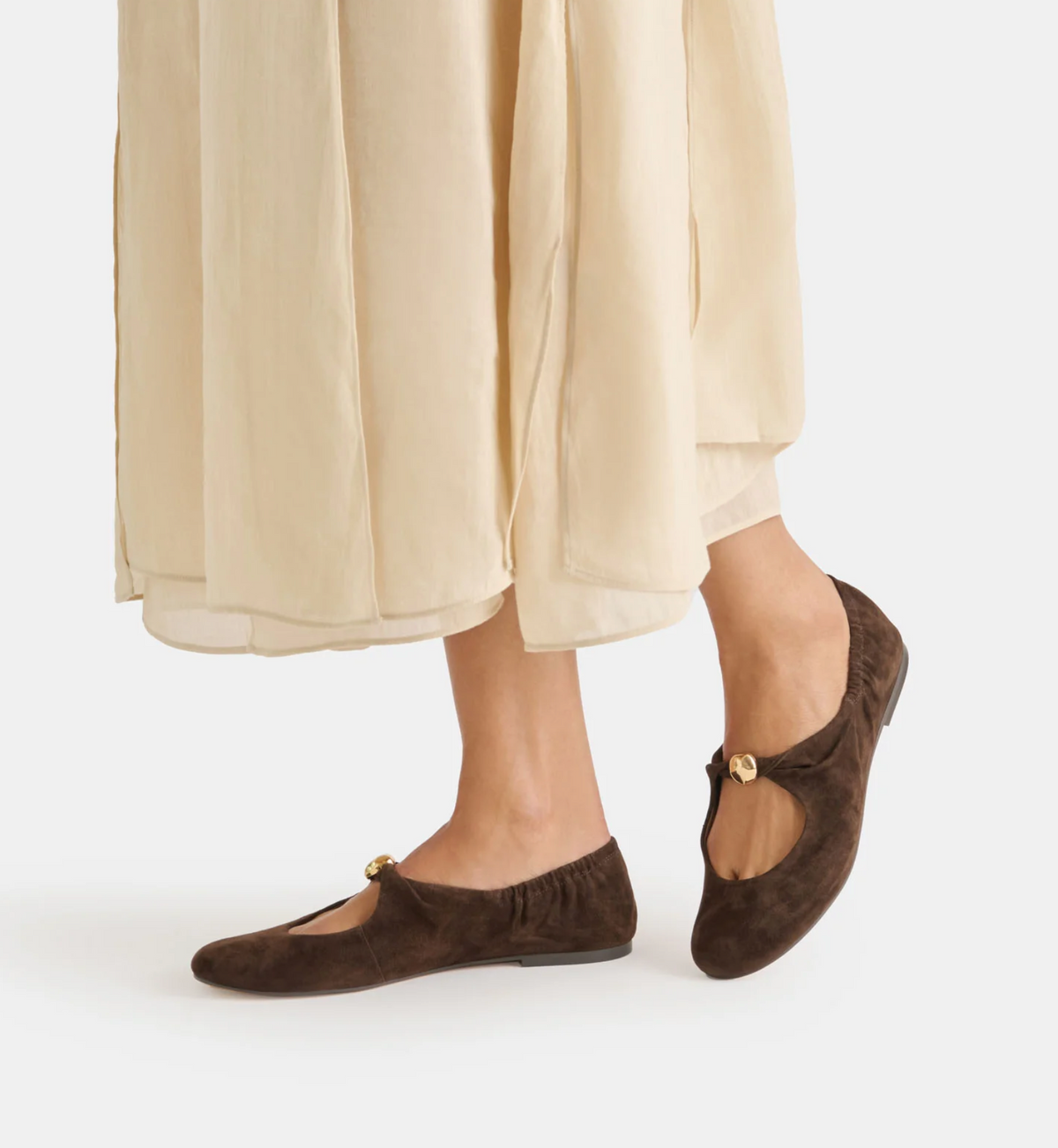 A person wearing dark brown Dolce Vita Cornel suede Mary Jane flats with a small gold button detail, paired with a flowing cream-colored skirt, shown from the knees down against a light gray studio background.