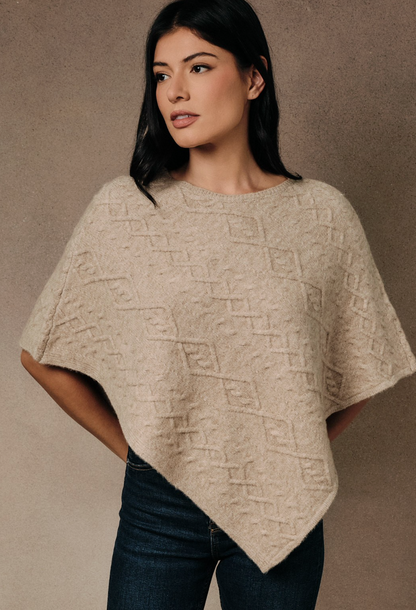 A woman wearing a beige textured knit poncho sweater with a geometric pattern, paired with dark blue jeans, against a neutral textured studio backdrop.