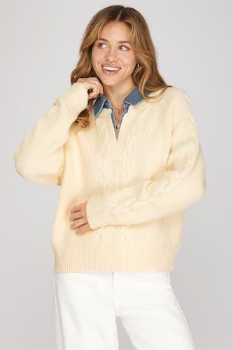 A woman wearing a cream-colored cable-knit sweater over a blue collared shirt, paired with white pants, against a solid light gray background.