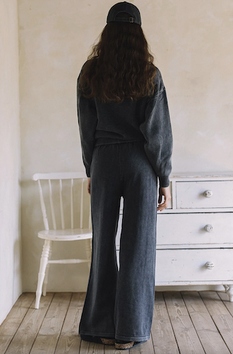 A woman stands with her back to the camera wearing a dark charcoal gray matching lounge set consisting of a long-sleeve sweatshirt and wide-leg pants, paired with a black baseball cap in a rustic room with a white dresser and wooden floor.