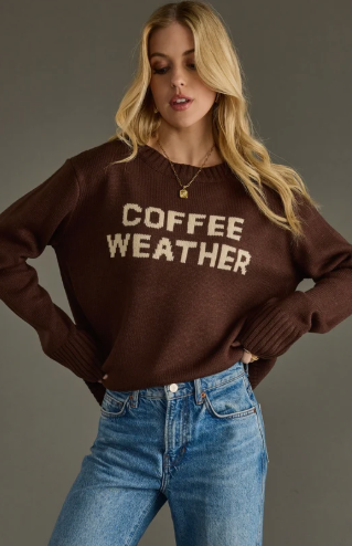 A woman wearing a brown crewneck sweater with 'COFFEE WEATHER' written across the chest in cream lettering, paired with blue jeans, against a solid gray studio background.
