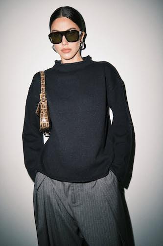 A model wearing a black mock-neck knit sweater and dark gray pinstripe trousers, accessorized with black sunglasses and a brown patterned shoulder bag, set against a plain light gray background.