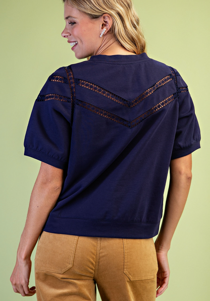 A woman shown from the back wearing a navy blue short-sleeve top with decorative crochet lace detailing across the shoulders and upper back, paired with tan pants, against a light green background.