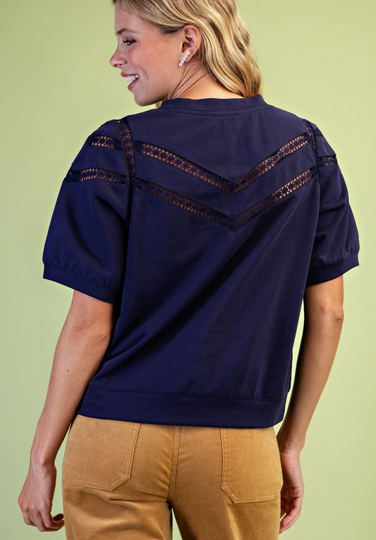 A woman shown from the back wearing a navy blue short-sleeve top with decorative crochet lace detailing across the shoulders and upper back, paired with tan pants, against a light green background.