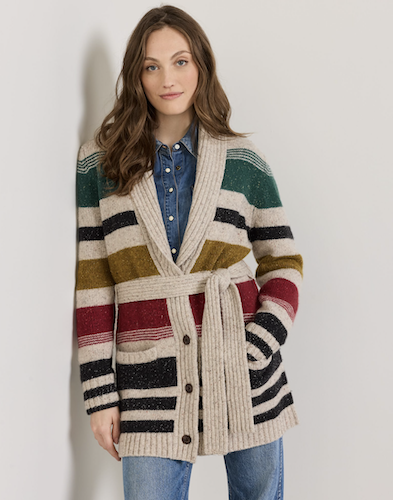 A woman models a Pendleton chunky wool-knit cardigan with horizontal stripes in beige, black, green, mustard yellow, and red, worn over a blue denim shirt and jeans against a plain light gray background.