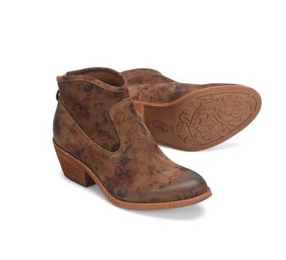 A pair of Sofft Aisley brown suede ankle boots with a distressed floral pattern, featuring a low block heel and a side cutout. One boot is shown from the side profile, and the other is positioned behind it to display the embossed floral sole.