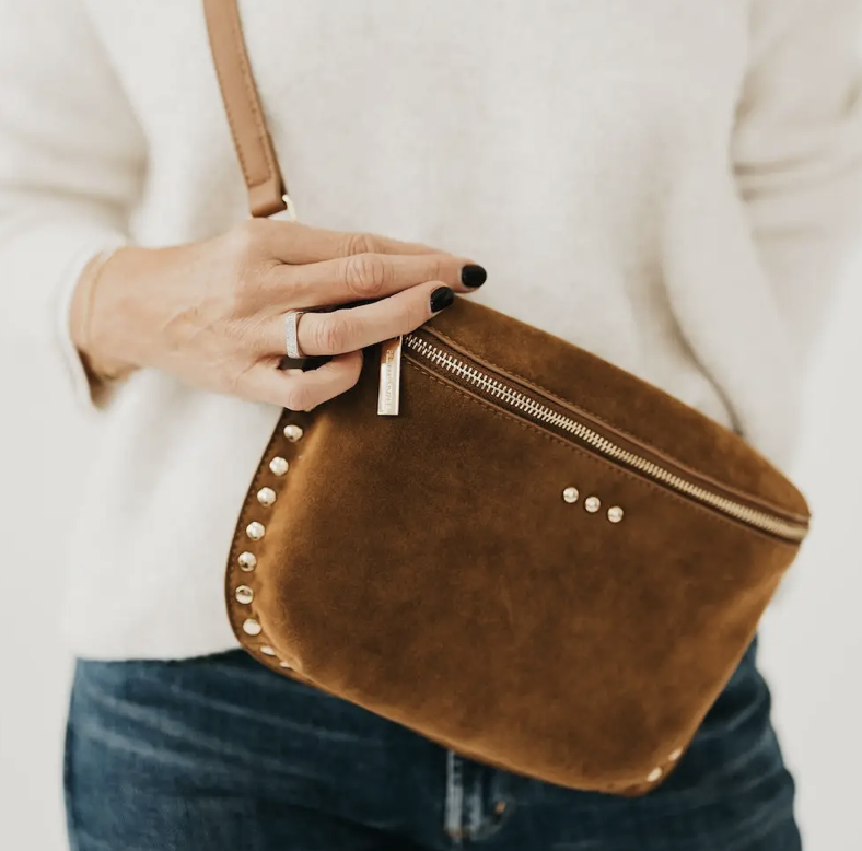 A person wearing a cream sweater and blue jeans holds a brown suede crossbody bag with gold-tone studs and a zipper.