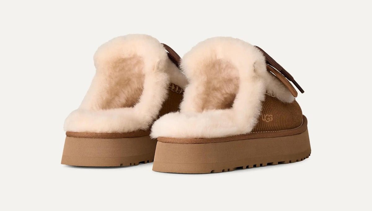 A pair of UGG platform Disquette Heritage slippers with thick tan soles and fluffy cream shearling lining, shown from a three-quarters angle on a light off-white background.