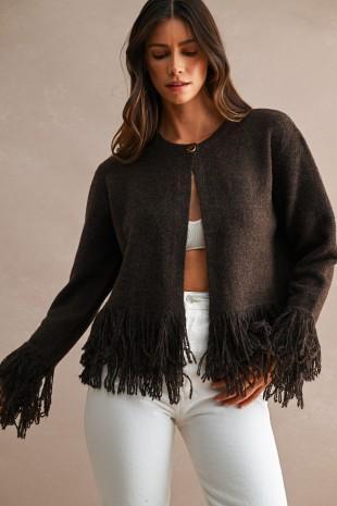 A woman wearing a dark brown open-front knit cardigan with long fringe trim along the hem and cuffs, paired with white pants, against a neutral beige studio background.