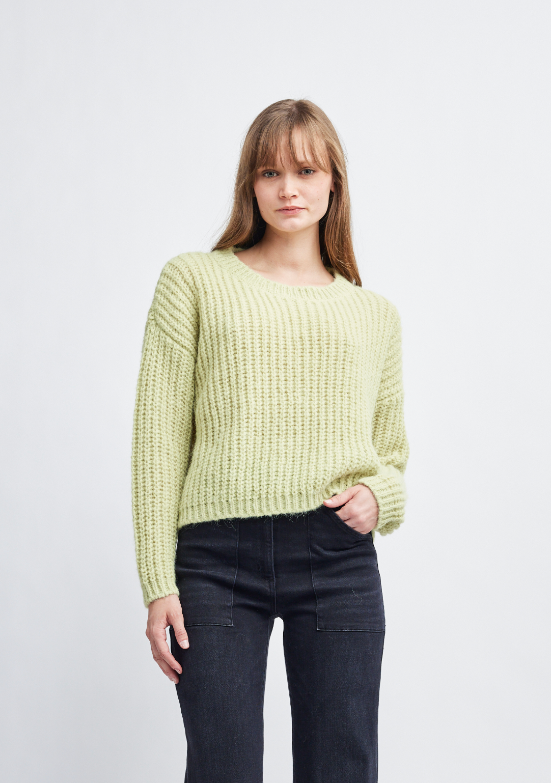 A woman modeling a light yellow-green chunky knit crewneck sweater paired with dark jeans, standing against a plain light gray studio background.