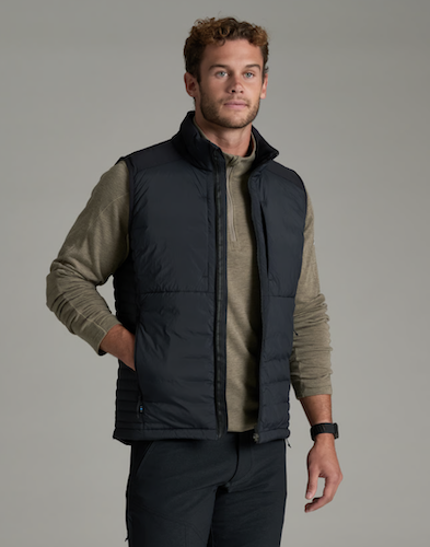 A man wearing a black Kuhl Spyfire quilted puffer vest over a beige long-sleeve shirt and dark pants, standing against a solid gray studio background.
