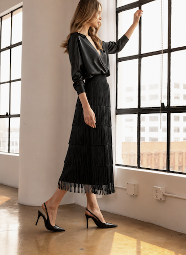 A woman models a black satin blouse and a black pleated midi skirt with fringe, paired with black pointed-toe heels, in a bright loft setting with large industrial windows.