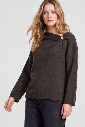 A woman wearing a dark brown knit sweater with a unique asymmetrical draped neckline and a gold-tone decorative brooch ring detail, paired with dark jeans against a light gray background.