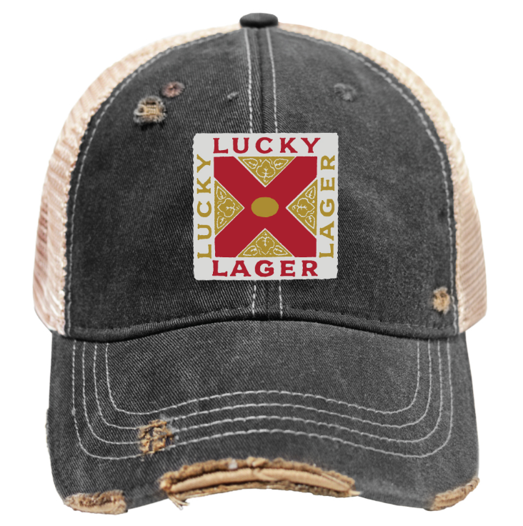 A distressed trucker-style baseball cap with a dark gray washed front and brim, beige mesh back panels, and a square embroidered patch on the front reading 'LUCKY LAGER' with a red and gold cross design.