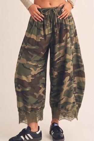 A person wearing loose camouflage barrel pants with a drawstring waist and lace trim at the ankles, paired with black sneakers, against a plain light beige background.