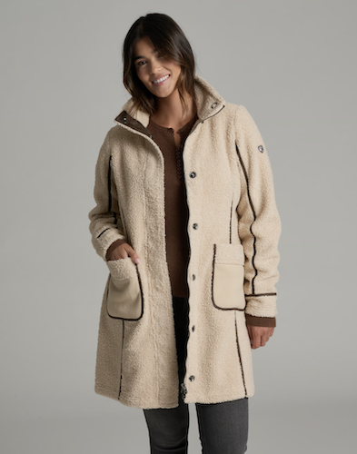 A woman wearing a beige Kuhl Durango sherpa fleece coat with brown trim over a brown top and dark gray pants, photographed against a solid gray studio background.