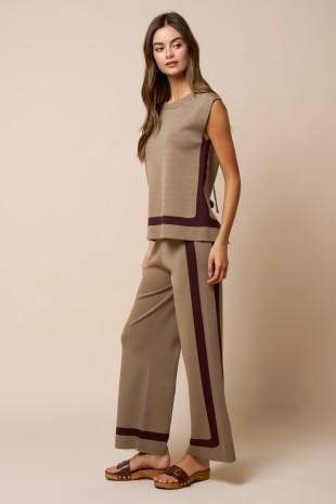 A woman modeling a sleeveless knit top and matching wide-leg pants set in taupe with dark plum side panels and trim, paired with brown sandals, against a solid beige studio background.