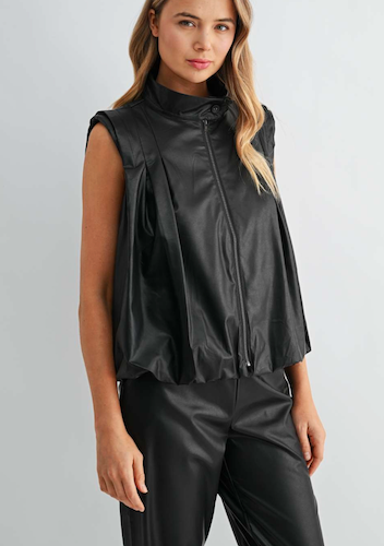 A woman wearing a sleeveless black faux-leather vest with a high mock neck and front zipper, paired with matching black pants, against a light gray studio background.