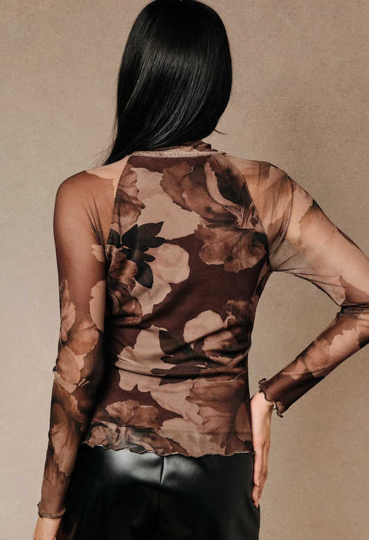 A woman shown from the back wearing a sheer long-sleeve top with a brown and beige floral print, paired with black pants, against a neutral textured beige background.