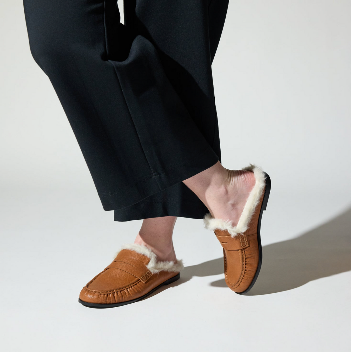A person wearing black wide-leg trousers and Yosi Samra Camden brown slip-on loafers with white shearling lining, shown from the calves down against a clean white studio background.