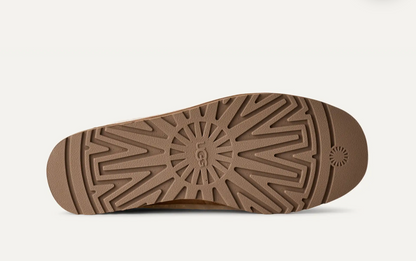 The brown rubber outsole of a UGG shoe, featuring a textured radial tread pattern and the embossed UGG logo in the center, shown against a light off-white background.